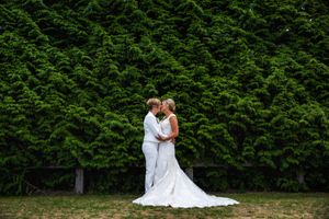 Two Brides kissing in front of tall hedge